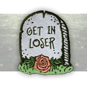 May include: Embroidered patch in the shape of a tombstone with the words "GET IN LOSER" in black. A pink rose with green leaves sits at the bottom, above a patch of green grass. The patch has a white background.