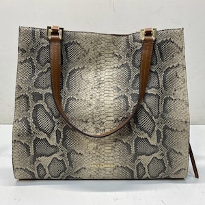 May include: A square-shaped tote bag featuring a grey and black snakeskin pattern. The bag has two brown leather shoulder straps attached with gold-tone metal hardware. The brand name VINCE CAMUTO is printed in small gold letters on the front.