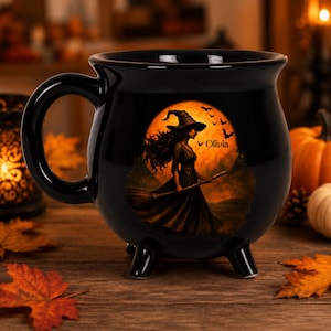 May include: A black ceramic cauldron-shaped mug featuring a graphic of a witch in a pointed hat holding a broomstick against a large orange full moon. The name Olivia is printed in black text next to the witch, with silhouettes of flying bats.