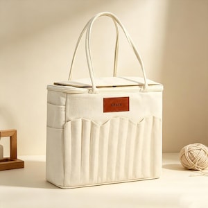 May include: A cream-coloured rectangular tote bag with two long handles, a zipped top closure, and side pockets. The front features a pleated panel and a brown rectangular leather-like patch with the word GRACE embossed in dark lettering.