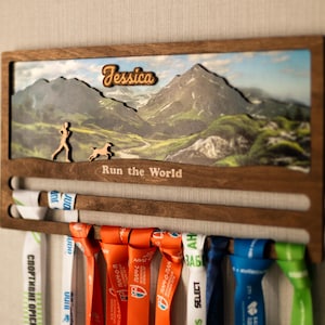 Puede incluir: Expositor de medallas de madera con una escena de montaña, el nombre "Jessica" y la frase "Run the World". El expositor tiene ranuras para medallas y siluetas de un corredor y un perro. Cintas de colores cuelgan del expositor.