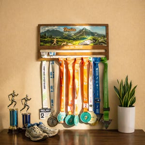 Könnte beinhalten: Ein hölzernes Medaillen-Display mit einem Berglandschaftsdruck, dem Namen Jessica und dem Text Run the World. Es hält mehrere bunte Bänder mit Medaillen, begleitet von zwei Läufer-Trophäen und einem Paar Sportschuhe.