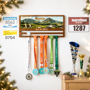 Puede incluir: Un expositor de medallas de madera con un estampado de paisaje montañoso, el nombre Cameron y el texto Run the World. Tiene dos barras horizontales para colgar medallas en cintas de colores, acompañado de tres dorsales de carrera y dos trofeos.