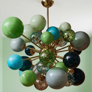 May include: A mid-century modern chandelier with a brass frame and multiple spherical glass globes in shades of green, blue, and gray. The light fixture is suspended from the ceiling, showcasing a unique and artistic design. The globes vary in size and texture.
