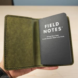 May include: An olive green leather notebook cover with black stitching, holding a black Field Notes 48-Page Memo Book with white text that reads Field Notes, 48-Page Memo Book, Durable Materials / Made in the U.S.A. on the front cover.