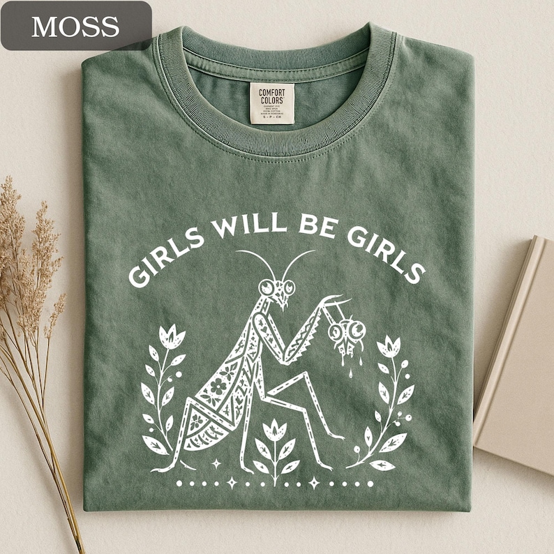 May include: Moss green t-shirt with the text "GIRLS WILL BE GIRLS" and a white graphic of a praying mantis holding a pair of eyeballs. The shirt has a crew neck and a Comfort Colors tag.