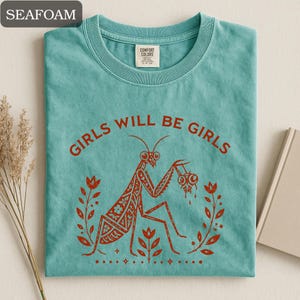 May include: Seafoam green t-shirt with the text "GIRLS WILL BE GIRLS" in red. The shirt features a red illustration of a praying mantis holding two eyeballs, surrounded by floral elements. The shirt is made of a soft material.