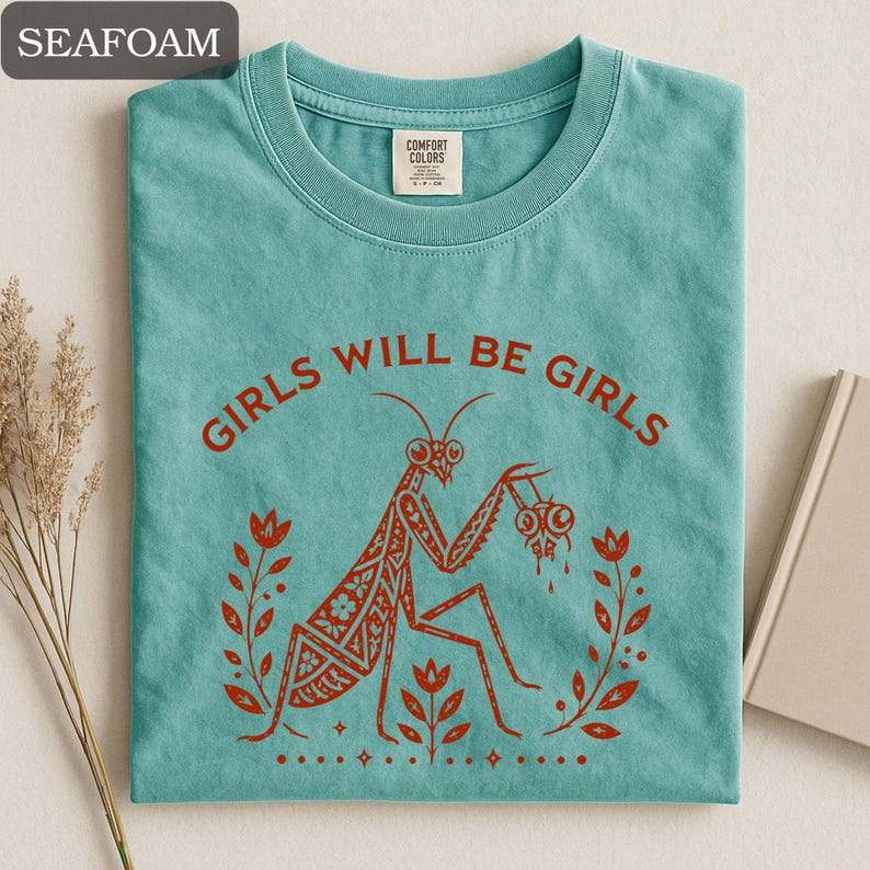 May include: Seafoam green t-shirt with the text "GIRLS WILL BE GIRLS" in red. The shirt features a red illustration of a praying mantis holding two eyeballs, surrounded by floral elements. The shirt is made of a soft material.