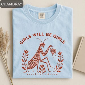 May include: Light blue t-shirt with the text "GIRLS WILL BE GIRLS" in a circular design. A detailed illustration of a praying mantis holding two objects is in the center, surrounded by floral elements. The shirt is made of a soft material.
