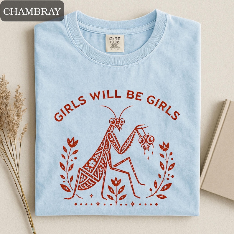 May include: Light blue t-shirt with the text "GIRLS WILL BE GIRLS" in a circular design. A detailed illustration of a praying mantis holding two objects is in the center, surrounded by floral elements. The shirt is made of a soft material.