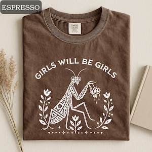 May include: Espresso-colored t-shirt with the text "GIRLS WILL BE GIRLS" in white. A white illustration of a praying mantis is centered below the text, surrounded by floral elements. The shirt has a crew neck and a Comfort Colors tag.