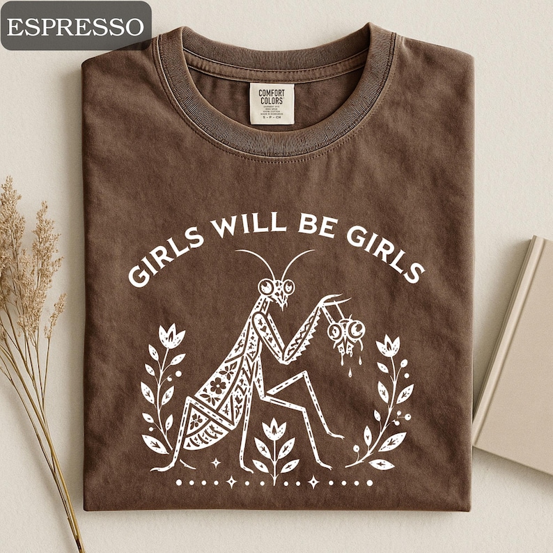 May include: Espresso-colored t-shirt with the text "GIRLS WILL BE GIRLS" in white. A white illustration of a praying mantis is centered below the text, surrounded by floral elements. The shirt has a crew neck and a Comfort Colors tag.