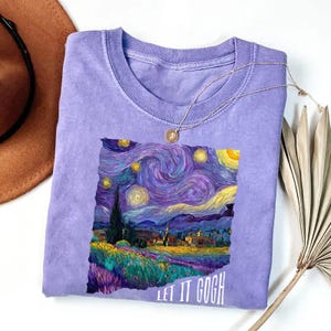 May include: A lilac-colored t-shirt featuring a print of Van Gogh's "Starry Night" painting. The artwork includes swirling blues, purples, and yellows, with the text "LET IT GOGH" in white. A silver necklace with a small pendant is draped across the shirt.