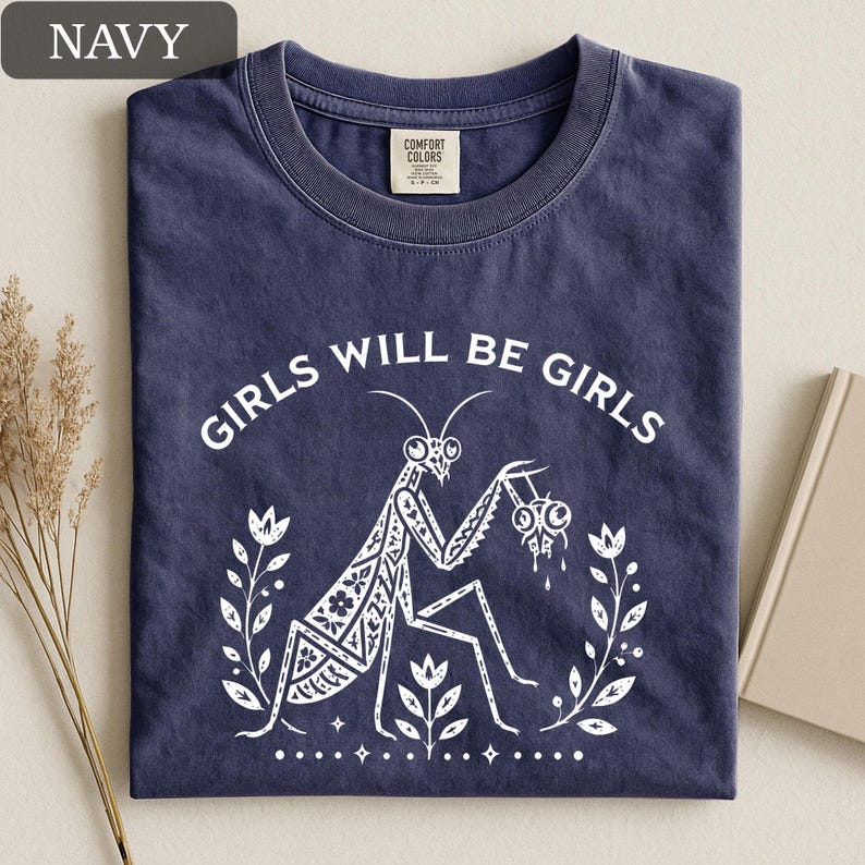 May include: Navy blue t-shirt with the text "GIRLS WILL BE GIRLS" and a white graphic of a praying mantis holding a pair of eyeballs. The design is surrounded by a floral wreath.