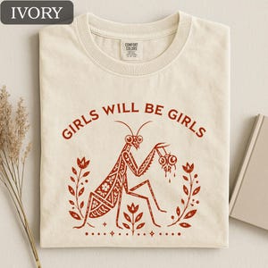 May include: Ivory-colored t-shirt with the text "GIRLS WILL BE GIRLS" in red. The shirt features a red illustration of a praying mantis holding eyeballs, surrounded by floral elements. The shirt is made of Comfort Colors material.