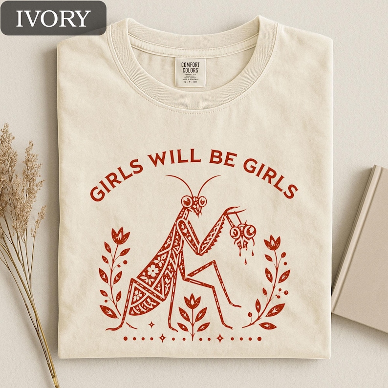 May include: Ivory-colored t-shirt with the text "GIRLS WILL BE GIRLS" in red. The shirt features a red illustration of a praying mantis holding eyeballs, surrounded by floral elements. The shirt is made of Comfort Colors material.