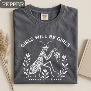 May include: A dark gray t-shirt with the text "GIRLS WILL BE GIRLS" in white. The shirt features a white graphic of a praying mantis holding a dripping object, surrounded by floral elements. The shirt is folded.
