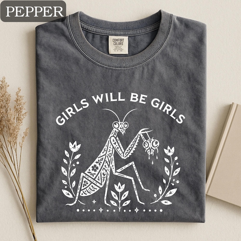 May include: A dark gray t-shirt with the text "GIRLS WILL BE GIRLS" in white. The shirt features a white graphic of a praying mantis holding a dripping object, surrounded by floral elements. The shirt is folded.