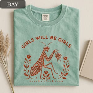 May include: A light green t-shirt with the text "GIRLS WILL BE GIRLS" in red. The shirt features a red illustration of a praying mantis holding two eyeballs, surrounded by floral elements.