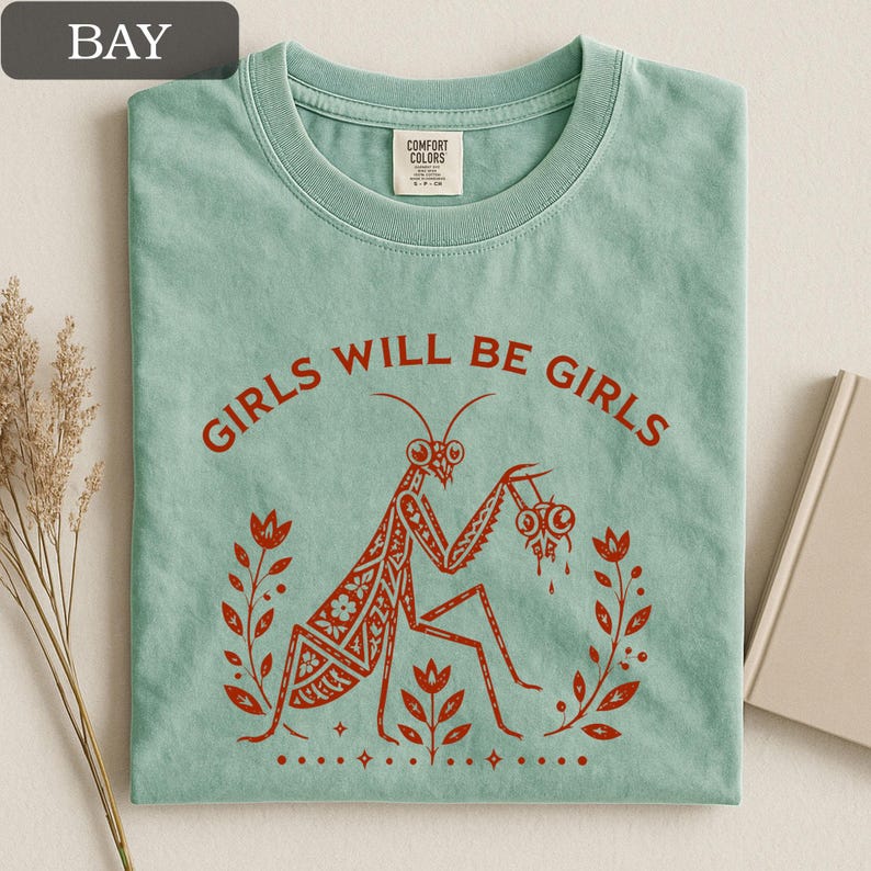May include: A light green t-shirt with the text "GIRLS WILL BE GIRLS" in red. The shirt features a red illustration of a praying mantis holding two eyeballs, surrounded by floral elements.