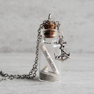 May include: A silver chain necklace with a small glass bottle pendant. The bottle contains sand, a seashell, and a rolled-up piece of paper with the text "Your Text Here". A silver anchor charm hangs from the bottle.