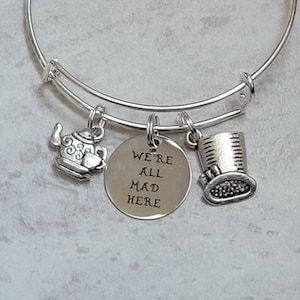 May include: Silver-toned bangle bracelet with three charms. Charms include a teapot, a top hat, and a circular charm with the text "WE'RE ALL MAD HERE". The bracelet is a whimsical accessory.