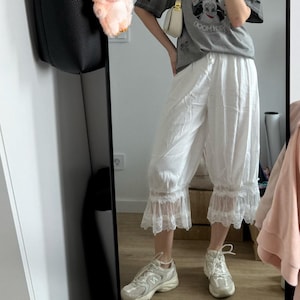 May include: A gray t-shirt with a graphic of three cartoon characters, paired with white capri pants with lace trim. A black tote bag hangs on the left, and a small plush toy is attached to it. The person is wearing white sneakers.