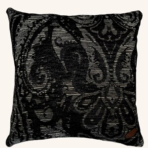May include: Square decorative pillow with a black and gray paisley pattern. The pillow has a textured appearance and a dark border. A small brown tag is sewn on the lower right corner.