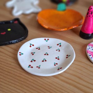May include: A small, white ceramic dish with a hand-painted cherry pattern. The dish is oval-shaped and features red cherries with green stems. Other decorative ceramic pieces are in the background, including a black cat, a ghost, and a pumpkin.