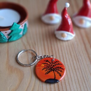 Peut inclure: Un porte-clés rond avec un motif de coucher de soleil représentant une silhouette d'arbre noir sur un ciel orange et jaune. Le porte-clés a un anneau et une chaîne argentés. En arrière-plan, de petits gnomes rouges et blancs et un bougeoir.
