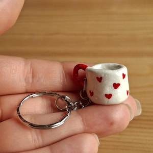 Pode incluir: Um pequeno porta-chaves de caneca branca com detalhes de corações vermelhos e uma alça vermelha. O porta-chaves está preso a um anel de metal prateado. A caneca é segurada em uma mão contra um fundo de madeira.