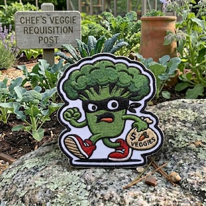 May include: An embroidered patch featuring a cartoon broccoli character wearing a black eye mask, running while holding a tan sack labeled with a dollar sign and the text VEGGIES. The character has green skin, red and white sneakers, and a determined expression.