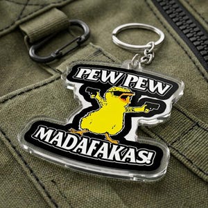 May include: A clear acrylic keychain featuring a yellow chick wearing sunglasses and holding two guns. The keychain has the words "PEW PEW" and "MADAF*KAS!" in white text on a black background. The keychain is attached to a silver keyring.