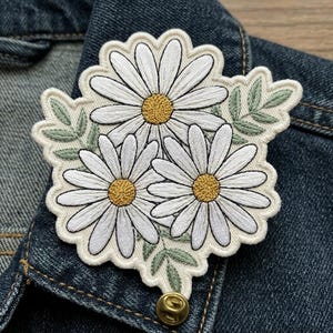 May include: Embroidered daisy patch featuring three white daisies with yellow centers and green leaves. The patch is attached to a denim jacket with a gold-colored pin.