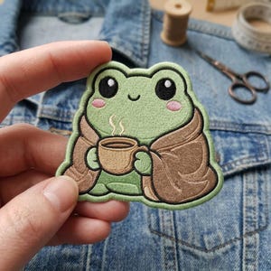 May include: Embroidered patch featuring a cartoon frog in a brown blanket, holding a coffee cup. The frog is light green with pink cheeks and black eyes. Steam rises from the cup. The patch is likely for clothing or accessories.