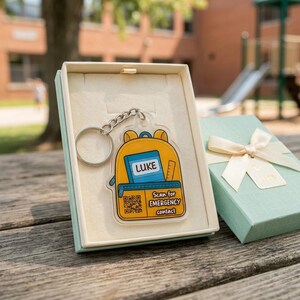 May include: A clear acrylic keychain featuring a yellow backpack design with the name "LUKE" on a blue book. The keychain includes a QR code and the text "Scan for EMERGENCY contact". It is presented in a light blue gift box.
