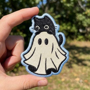 May include: An embroidered patch featuring a black cat wearing a white ghost costume. The cat has large white eyes and is peeking out from the top of the sheet. The ghost costume has two black eye holes and a light blue border around the edge.