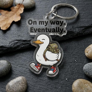 May include: A clear acrylic keychain featuring a cartoon duck wearing red sneakers and a backpack. The keychain has the text "On my way. Eventually." in black. The duck has a yellow beak and is smiling. A silver key ring is attached.