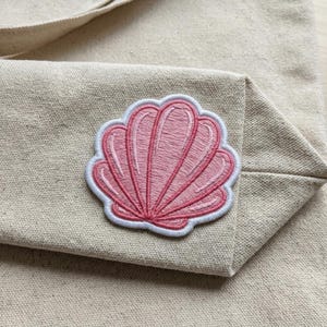 May include: A pink seashell patch with white trim is sewn onto a beige canvas tote bag. The embroidered seashell design features detailed stitching and a vibrant pink color, adding a decorative element to the bag. The tote bag is made of a sturdy canvas material.
