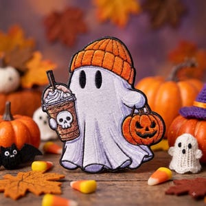 May include: An embroidered patch featuring a white ghost wearing an orange beanie, holding a brown iced coffee cup with a skull graphic in one hand and a small orange jack-o'-lantern pumpkin bucket in the other hand, with black outlines.