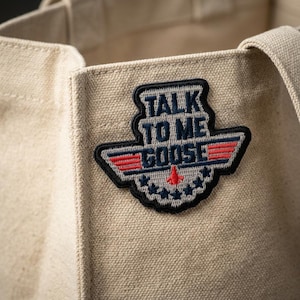 May include: Beige canvas tote bag with a patch that reads "TALK TO ME GOOSE" in white and blue lettering. The patch has a red and blue wing design and a small red airplane. The bag has a simple design with a handle.