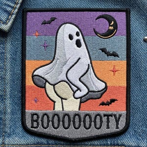May include: Embroidered patch featuring a cartoon ghost with the word "BOOOOOOTY". The patch has a colorful background with bats, stars, and a crescent moon. The ghost is white with black eyes and mouth.