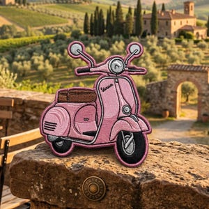 May include: An embroidered patch shaped like a pink Vespa scooter with a brown seat, black tires, and silver handlebars. The patch features the Vespa logo in black text on the front panel and is designed with detailed stitching throughout the body.