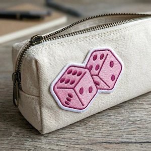 May include: A beige canvas pencil case with a bronze zipper and a pink dice patch. The dice patch features two dice with pink dots and a white border. The pencil case is rectangular and has a zipper closure.