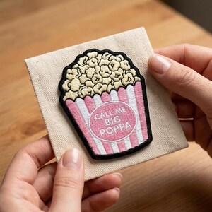 Puede incluir: Parche bordado con un diseño de cubo de palomitas de maíz con rayas rosas y blancas, lleno de palomitas de maíz. El texto "CALL ME BIG POPPA" está en un círculo rosa. El parche está sobre una tela beige.