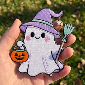 May include: An embroidered patch featuring a white ghost wearing a lavender witch hat with a mint green band and a small dangling star. The ghost has pink cheeks, holds an orange jack-o'-lantern bucket, and carries a mint green broomstick.