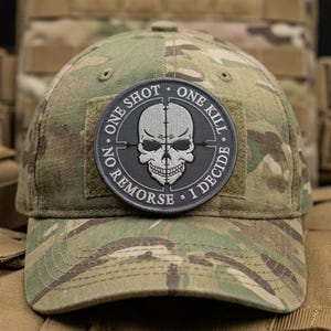 May include: Camouflage baseball cap with a circular patch featuring a skull with crosshairs and the text "ONE SHOT ONE KILL NO REMORSE I DECIDE". The cap has a curved brim and a structured crown.