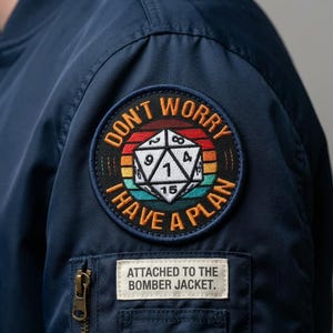 May include: Navy blue bomber jacket with a circular patch featuring a D20 die and the text "DON'T WORRY, I HAVE A PLAN." A smaller rectangular patch below reads "ATTACHED TO THE BOMBER JACKET."