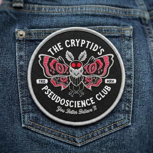 May include: A round black patch with white stitching, featuring a moth design in red and white, and the text "THE CRYPTID'S PSEUDOSCIENCE CLUB". The phrase "You Better Believe It" is also included. The patch is sewn onto a blue denim pocket.