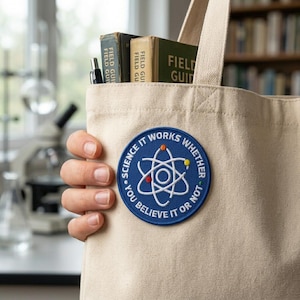 May include: A beige canvas tote bag with a blue circular patch that reads "SCIENCE IT WORKS WHETHER YOU BELIEVE IT OR NOT." The bag contains vintage books and a pen. The patch features a white atom design with red, yellow, and blue accents.